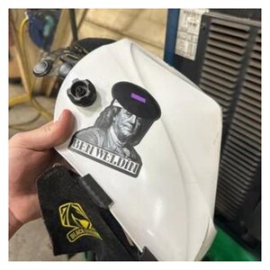 May include: White welding helmet with a black and purple lens. The helmet features a sticker with an image of Benjamin Franklin and the text "BEN WELDIN". A black and yellow logo is visible on the bottom of the helmet.