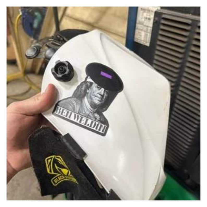 May include: White welding helmet with a black and purple lens. The helmet features a sticker with an image of Benjamin Franklin and the text "BEN WELDIN". A black and yellow logo is visible on the bottom of the helmet.