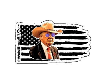 May include: A black and white distressed American flag with a portrait of a man wearing a cowboy hat and sunglasses. The text "WeldShop21.com" is on the left side of the image.