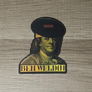 May include: A sticker featuring Benjamin Franklin wearing a welding helmet. The sticker has a gold and black color scheme with the words "BEN WELDIN" in a gold, bold font. The image is on a wooden surface.