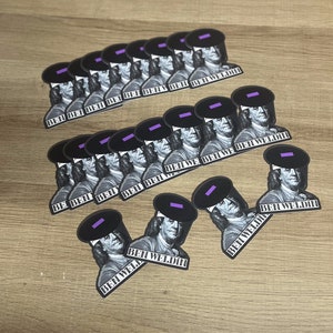 May include: A collection of stickers featuring a black and white image of Benjamin Franklin wearing a welding helmet. The stickers have the text "BEN WELDIN" printed on them. The stickers are arranged on a light brown wooden surface.