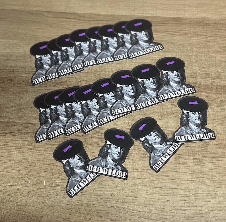 May include: A collection of stickers featuring a black and white image of Benjamin Franklin wearing a welding helmet. The stickers have the text "BEN WELDIN" printed on them. The stickers are arranged on a light brown wooden surface.