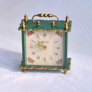 May include: A square, green Intertime clock with gold-coloured accents. The clock face is cream-coloured with Roman numerals and small floral accents. A gold handle sits atop the clock.