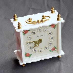 May include: A white decorative clock with gold accents and a handle. The clock face features Roman numerals, red rose accents, and the brand name "Jensor". The clock is approximately 4 inches tall.