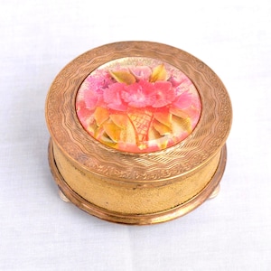 May include: A round gold-tone metal powder compact featuring a textured, engraved border on the lid. The centre of the lid displays a circular inlay with a colourful floral design of pink and red flowers with green leaves against a light background.