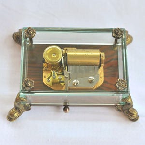 May include: A vintage music box with a glass case and brass accents. The box features a visible mechanical movement with gold-colored components and a wooden base. The corners and feet have ornate brass detailing, adding to its antique charm.