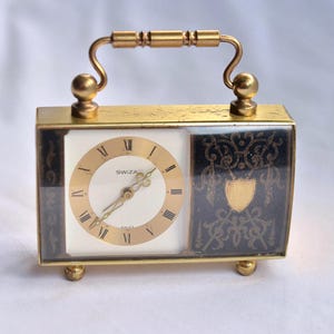 May include: A vintage gold-coloured travel clock with a handle. The clock face is white with gold hands and Roman numerals. The sides are black with gold decorative patterns. The clock has a rectangular shape and sits on small, rounded feet.