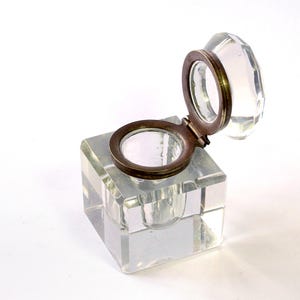 May include: A clear glass inkwell featuring a cube-shaped base and a hinged circular lid. The lid is constructed from a metallic bronze-toned frame with a faceted glass insert. The inkwell has a central circular opening for ink storage.