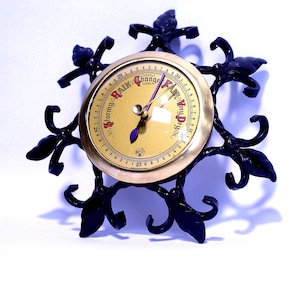 May include: A circular barometer with a gold-toned face featuring black text reading Stormy, Rain, Change, Fair, and Very Dry. The device has two dark purple hands and is encased in a black metal frame with decorative leaf-like scrollwork.