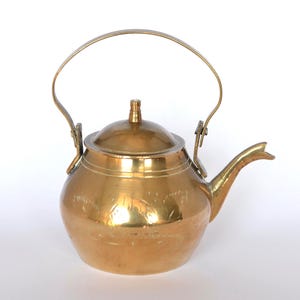May include: A polished brass tea kettle featuring a rounded body, a curved spout, and a hinged arched handle. The lid is domed with a small central knob, and the surface of the kettle displays subtle etched floral patterns around the midsection.