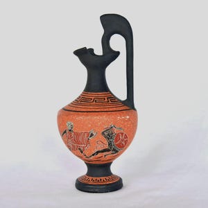 May include: An orange and black ceramic vase with a handle and spout. The vase features a black base and neck, with a decorative band around the neck. The body of the vase depicts figures in red and black, possibly from Greek mythology.