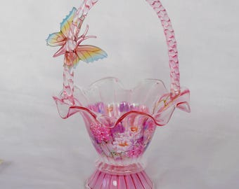 May include: A pink glass basket with a handle and a butterfly accent. The basket features a ruffled rim and a floral design on the interior. The handle is braided, and the butterfly has yellow, blue, and pink wings.
