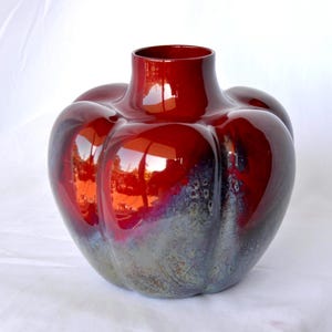 May include: A glossy, red art glass vase with a unique, rounded shape. The vase has a wide base that tapers to a narrow neck. The color transitions from a deep red at the top to a gray-blue at the bottom, creating a striking visual effect. A decorative piece.
