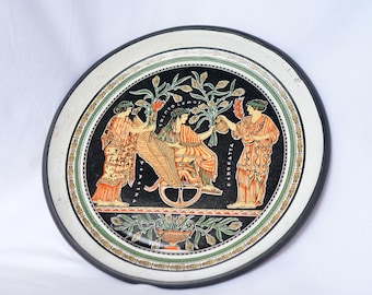 May include: A round decorative plate featuring a black center with a classical Greek-style illustration of three figures, including one seated in a winged chariot. The text TRINTOLEMOS and PEROFATTA is visible. The border has green and gold patterns.
