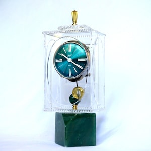 Puede incluir: Un reloj de sobremesa con esfera circular de color verde azulado, números romanos y manecillas blancas, encerrado en una caja rectangular de acrílico transparente con un remate dorado. El reloj descansa sobre una base de piedra verde oscuro.