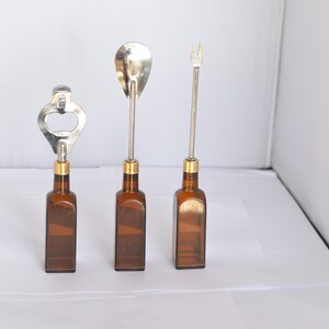 May include: Three bar tools: a bottle opener, a spoon, and a fork. Each tool has a silver metal top and a brown glass bottle base. The bottle opener has a silver metal top with a gold-colored band. The spoon and fork have a gold-colored band.