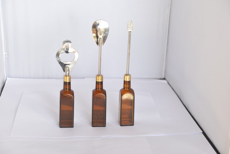 May include: Three bar tools: a bottle opener, a spoon, and a fork. Each tool has a silver metal top and a brown glass bottle base. The bottle opener has a silver metal top with a gold-colored band. The spoon and fork have a gold-colored band.