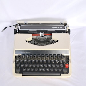 May include: Cream-colored Adler Tippa de Luxe typewriter with black keys and a red tab key. The typewriter has a silver trim and the text "ADLER" is visible. The carriage return lever is extended.