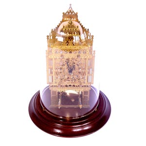 May include: A gold-coloured metal skeleton clock featuring intricate architectural detailing and a circular clock face with black hands, enclosed within a clear glass dome that rests upon a circular, dark-stained wooden base.