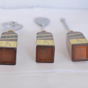 May include: Three bar tools with brown rectangular handles and metal tops. The tools include a bottle opener, a spoon, and a stirrer. The handles have labels with text and a logo. The tools are arranged on a white surface.