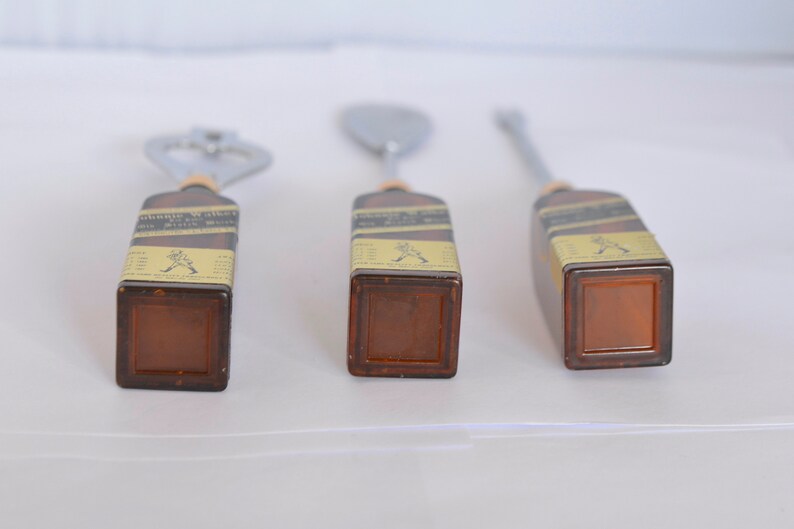 May include: Three bar tools with brown rectangular handles and metal tops. The tools include a bottle opener, a spoon, and a stirrer. The handles have labels with text and a logo. The tools are arranged on a white surface.