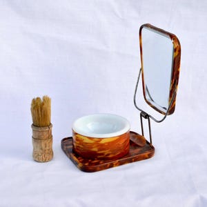 Könnte beinhalten: Ein Vintage-Rasier-Set mit Pinsel, Schale und Spiegel. Schale und Spiegelrahmen weisen ein Schildpattmuster auf. Der Pinsel hat einen Holzgriff und Borsten. Das Set wird vor weißem Hintergrund präsentiert.
