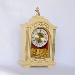 Puede incluir: Reloj de sobremesa Seiko color crema con detalles dorados y una pequeña figura de ángel en la parte superior. La esfera es dorada con números romanos negros y fondo rojo. El péndulo es visible.