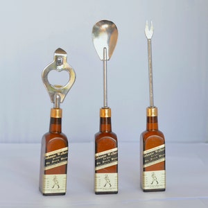 May include: Three bar tools with metal tops attached to miniature brown glass bottles. The tools include a bottle opener, a spoon, and a fork. Each bottle has a label that reads "Johnnie Walker Old Scotch Whisky."