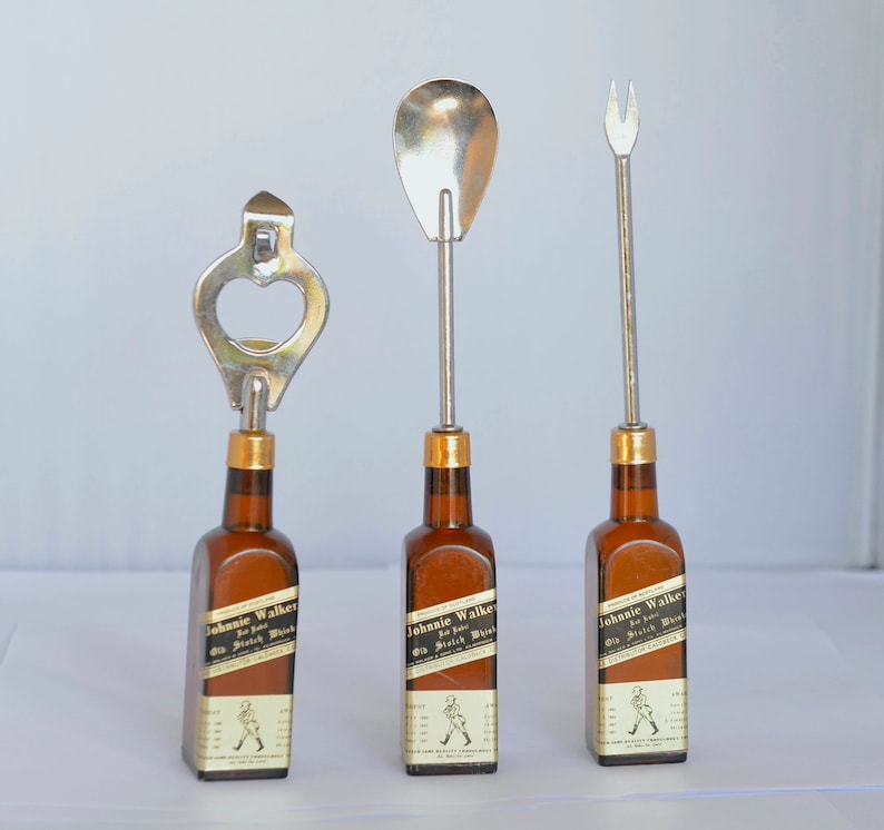 May include: Three bar tools with metal tops attached to miniature brown glass bottles. The tools include a bottle opener, a spoon, and a fork. Each bottle has a label that reads "Johnnie Walker Old Scotch Whisky."