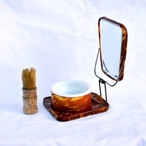 May include: A shaving set featuring a tortoiseshell-patterned stand with an attached rectangular mirror and a white ceramic bowl. Beside the stand is a wooden-handled shaving brush with light brown bristles, completing this grooming accessory set.