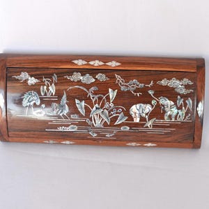 Puede incluir: Una caja rectangular de madera con acabado marrón oscuro, adornada con intrincados incrustaciones de nácar. La incrustación representa una escena serena con pájaros, elefantes y follaje. La caja tiene una parte superior curvada y es probable que se utilice para el almacenamiento.