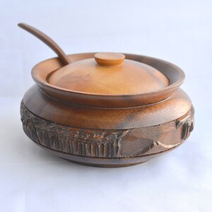 May include: A wooden sugar bowl with a lid and a spoon. The bowl is a warm brown color and has carved detailing around the base. The spoon rests on the rim of the bowl, which is also brown.