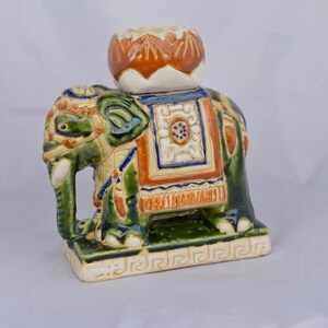 Puede incluir: Una figura de elefante de cerámica con un esmalte brillante en verde, naranja y azul. El elefante lleva un portavelas en forma de loto sobre su lomo y se apoya en una base rectangular decorada con un patrón geométrico de grecas.