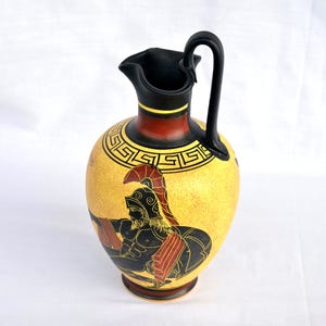 May include: A ceramic pitcher featuring a yellow body with a black and red painted illustration of a bearded warrior wearing a helmet. The neck is black with a yellow band, and the upper shoulder displays a black Greek key pattern design.