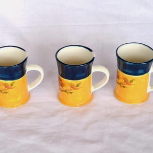 Puede incluir: Tres tazas de cerámica con acabado brillante, cada una con una sección superior azul marino profundo y una sección inferior amarilla brillante. La parte amarilla de cada taza está decorada con un pequeño patrón floral detallado en tonos naranja y verde.