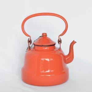 May include: A bright orange enamel kettle with a curved handle and spout. The kettle features a lid with a small chain and is set against a white backdrop. This retro kitchenware item is a classic design.
