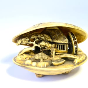 Könnte beinhalten: Eine cremefarbene, geschnitzte Muschelskulptur mit einer detaillierten Miniaturlandschaft im Inneren. Die Szene zeigt ein kleines Haus mit Strohdach, ein Wasserrad, einen Baum, eine Brücke und eine Figur neben einem Boot auf einem Sockel.