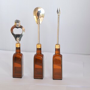 May include: Three bar tools: a bottle opener, a spoon, and a fork. Each tool has a silver metal top and a gold-colored band, attached to a brown glass bottle. The tools are arranged in a row against a white background.