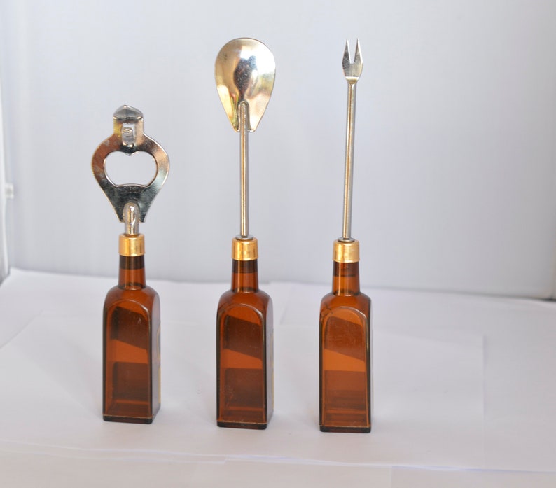May include: Three bar tools: a bottle opener, a spoon, and a fork. Each tool has a silver metal top and a gold-colored band, attached to a brown glass bottle. The tools are arranged in a row against a white background.