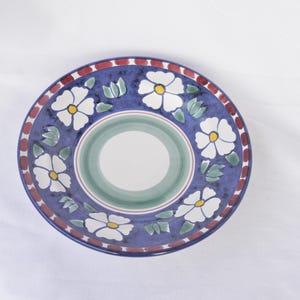 May include: A round, hand-painted ceramic plate with a blue background and white floral designs. The plate features a red and white border and a green and white centre. A decorative piece for the kitchen or dining room.