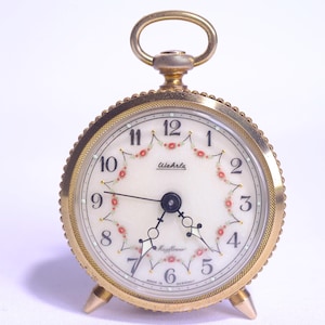 May include: A round gold-tone alarm clock with a white face featuring black Arabic numerals, ornate black hands, and a decorative floral garland pattern. The clock has a top handle, a textured bezel, and two small pointed feet at the base.