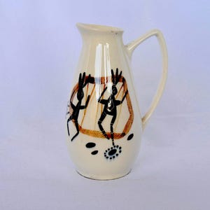 Puede incluir: Jarra de cerámica color crema con asa curva. La jarra presenta un diseño geométrico negro y marrón con figuras estilizadas. El diseño incluye una forma hexagonal marrón con figuras negras en el interior. La base tiene patrones de puntos negros y azules.