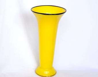 May include: A tall, bright yellow glass vase with a smooth, glossy finish. The vase features a wide, flared opening at the top with a thin, dark black rim that circles the entire circumference of the mouth, tapering down to a circular base.