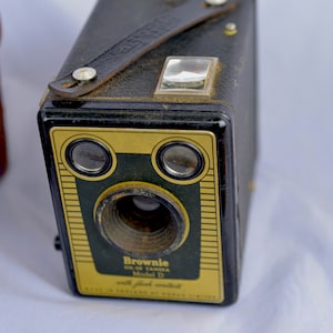 Puede incluir: Cámara vintage Brownie Model D, negra con detalles dorados y correa de cuero. La cámara tiene una lente con un anillo de metal y va acompañada de una funda de cuero marrón. La cámara está marcada "Brownie" y "Model D".