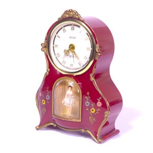 May include: A deep red mantel clock with gold-toned trim and floral accents. The clock face features Roman numerals and the brand name Peter. Below the dial, a glass-enclosed compartment contains a small figurine of a dancer in a white dress.