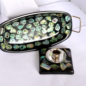 May include: An oval black tray with two gold-toned metal handles and an inlaid abalone shell mosaic pattern, accompanied by a square black coaster featuring a matching abalone shell inlay and a central silver-toned metal candle holder cup.