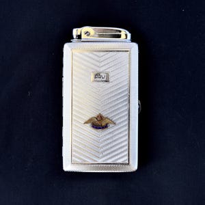 May include: A silver-toned cigarette case and lighter combination. The case has a textured, chevron-patterned front with the word "PAW" and a gold-toned emblem. The lighter is at the top, with a silver-toned body and a gold-toned top.