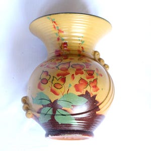 May include: A ceramic vase with a wide, ribbed neck and a rounded body. The base is dark brown, transitioning to a mustard yellow upper section. It features a painted floral design with orange-red petals and green leaves, plus side scroll accents.