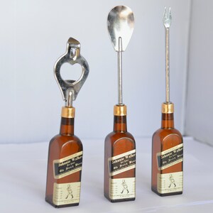 May include: Three bar tools: a bottle opener, a spoon, and a fork, each attached to a miniature brown glass bottle. The bottles have gold-colored caps and labels that read "Johnnie Walker Old Scotch Whisky".