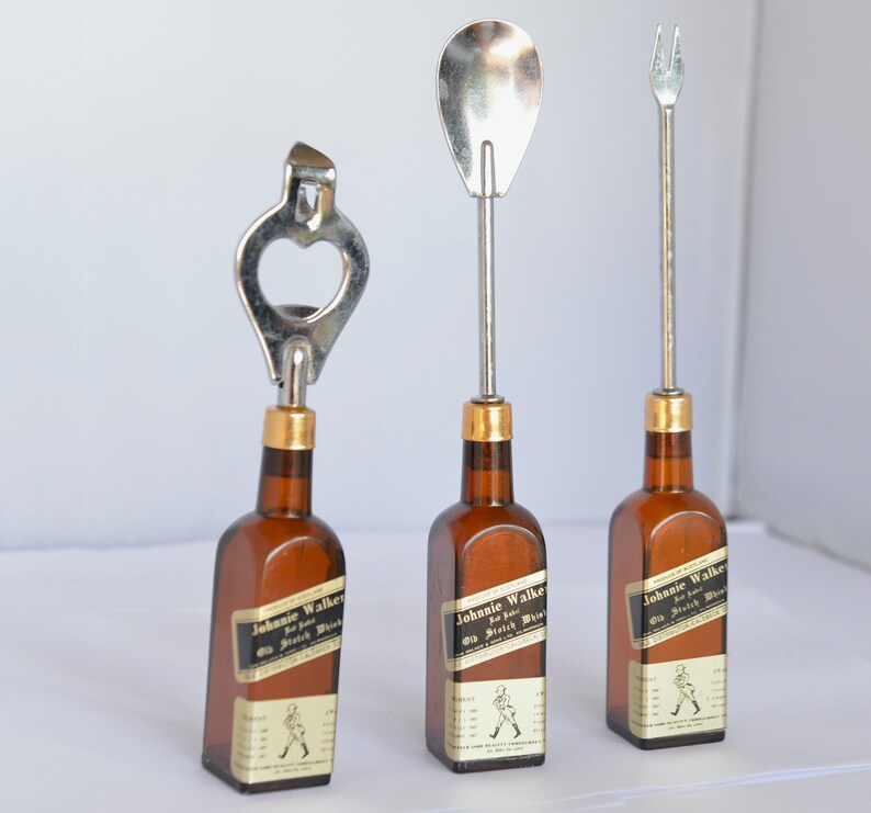 May include: Three bar tools: a bottle opener, a spoon, and a fork, each attached to a miniature brown glass bottle. The bottles have gold-colored caps and labels that read "Johnnie Walker Old Scotch Whisky".
